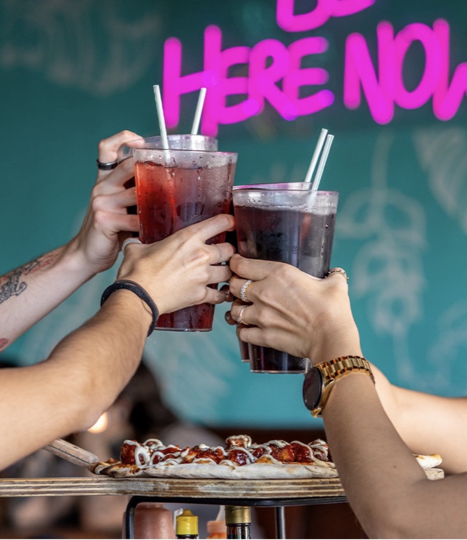 Brindis con bebidas bajo un neón rosa que dice Be here now